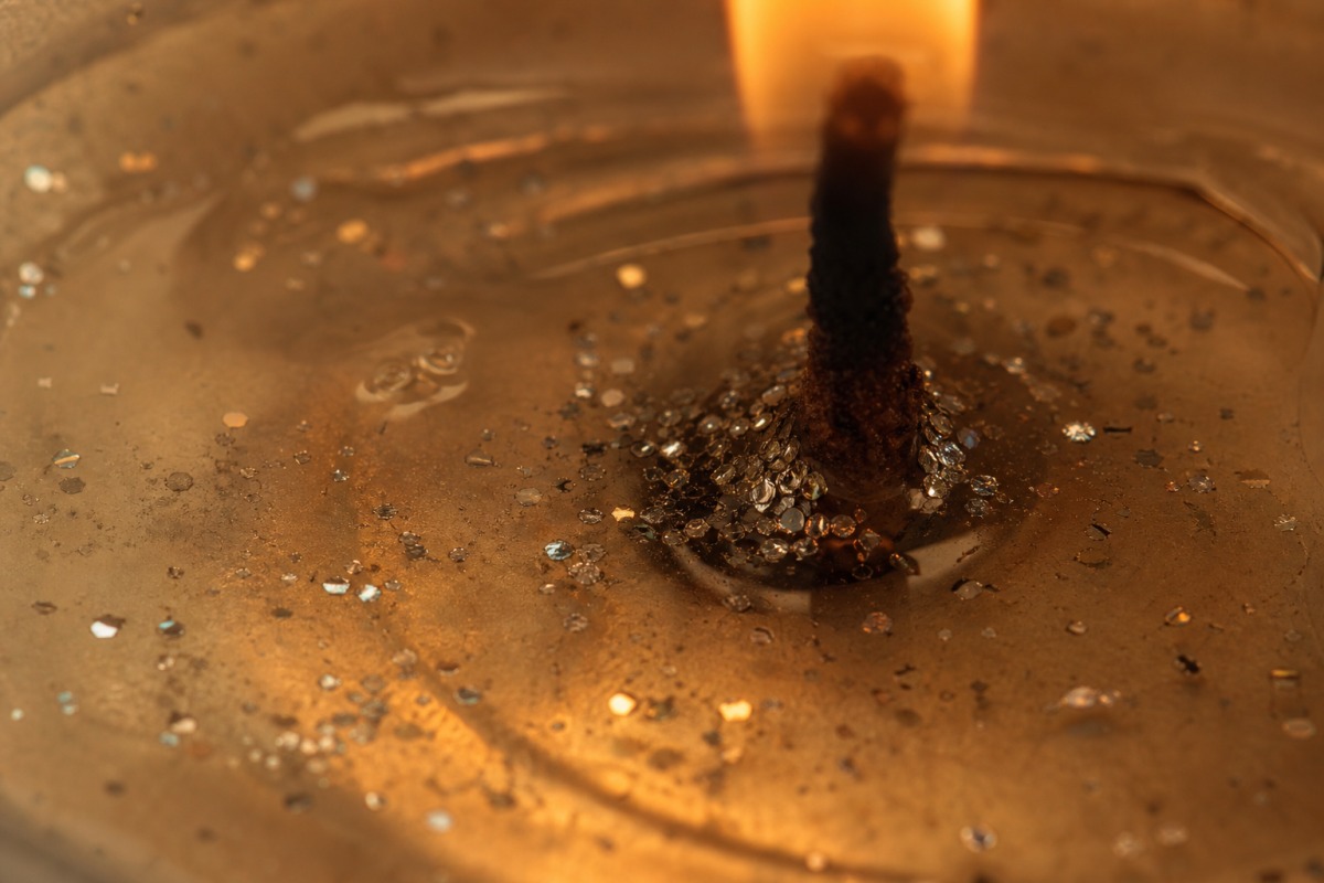 Macro de cera transparente derretida junto ao pavio de uma vela acesa, na qual descem minúsculos brilhantes prateados.