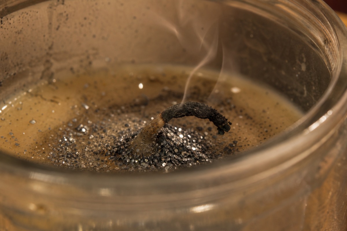 Pavio dobrado e carbonizado, coberto por um aglomerado de glitter prateado na cera solidificada, com a vela apagada.